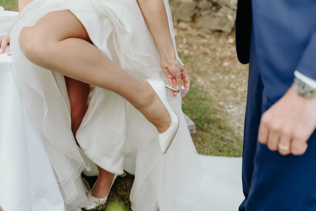 Dettaglio di una sposa che applica un salvatacco trasparente ai suoi tacchi bianchi prima di camminare sull’erba durante un matrimonio all’aperto