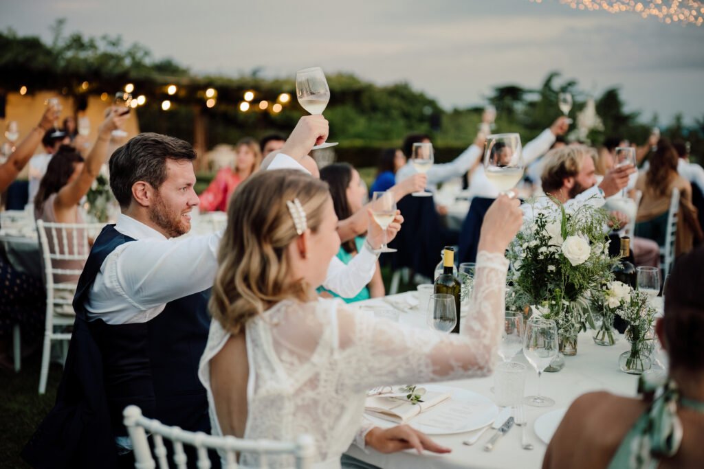 Ospiti alzano i calici durante un matrimonio elegante all’aperto, con tavoli decorati e luci sospese in un giardino al tramonto