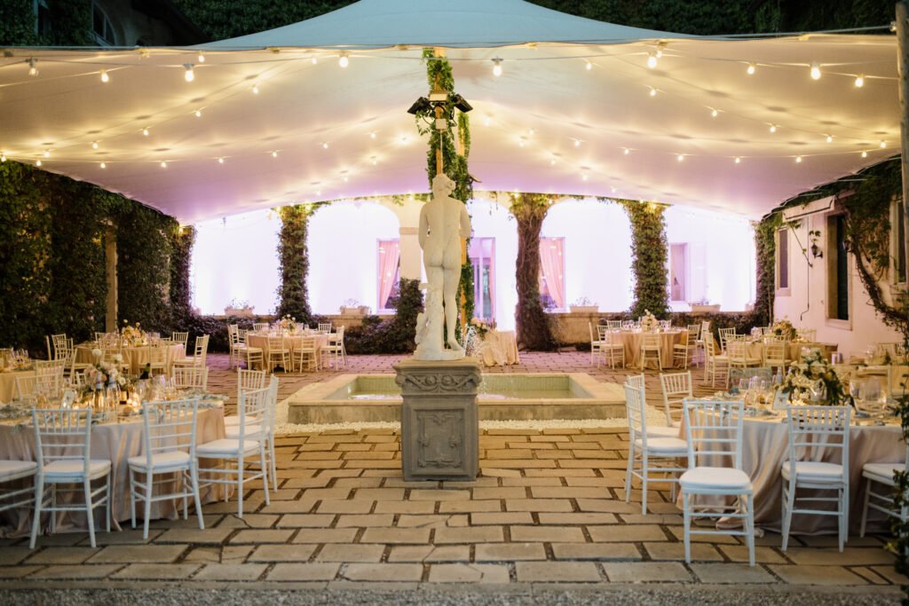 Allestimento da matrimonio serale sotto tensostruttura con luci calde, tavoli rotondi decorati, statue classiche e fontana centrale in una corte elegante
