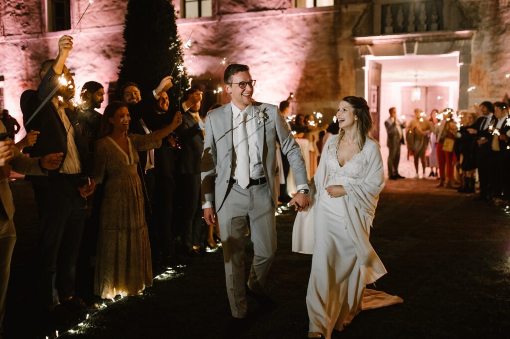 Sposi sorridenti camminano mano nella mano tra due ali di invitati che accendono sparkler durante la notte